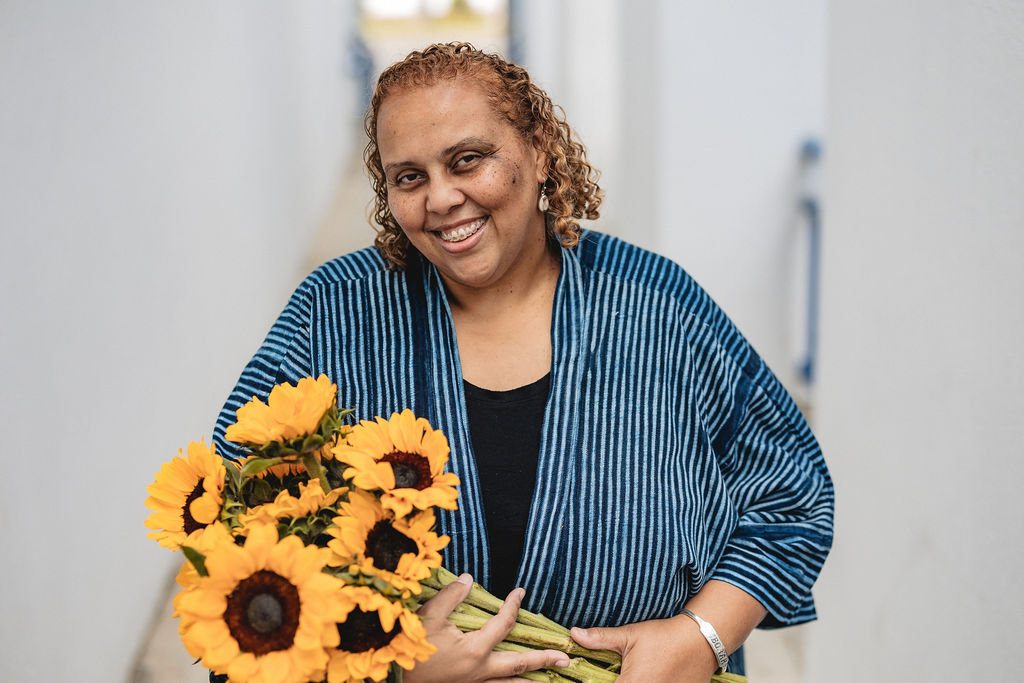 Deborah Anna Brown, smiling with curly hair holds a large bouquet of sunflowers. She is wearing a blue striped kimono style jacket and stands in a blurred bright white outdoor corridor.