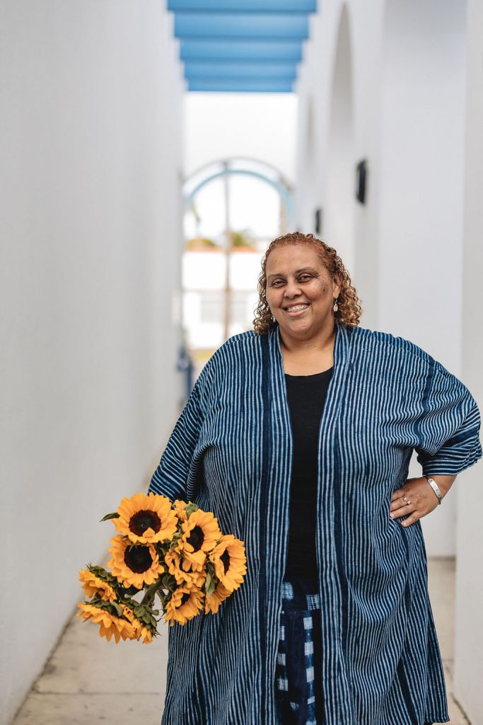 Deborah Anna Brown, smiling with curly hair stands in a blurred white outdoor hallway, holding a bouquet of sunflowers. She wears a blue striped kimono-style jacket.