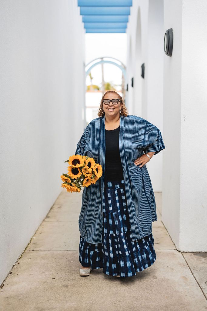 Deborah Anna Brown, smiling with curly hair and glasses stands in a white outdoor hallway, holding a bouquet of sunflowers. She wears a blue striped kimono-style jacket over a patterned blue skirt and off-white shoes.