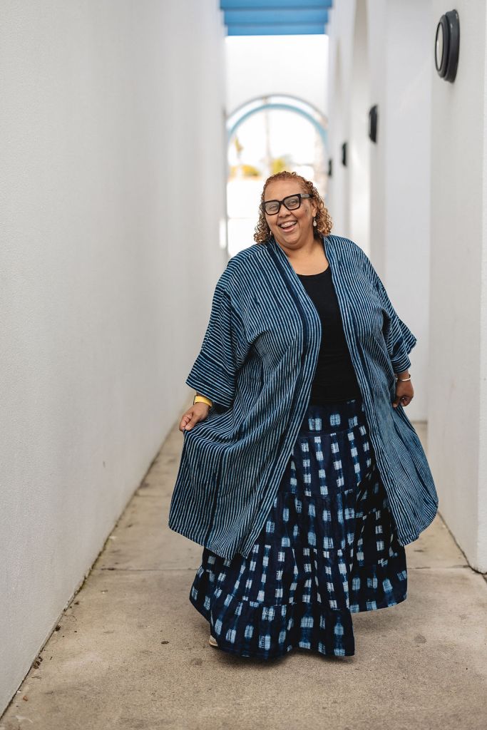 Deborah Anna Brown, smiling with curly hair and glasses in a blue striped kimono style jacket, dark shirt and patterned skirt are standing in a bright, white outdoor hallway with a blue slatted ceiling. She is holding her jacket, lifting it up and giving it a little frill.