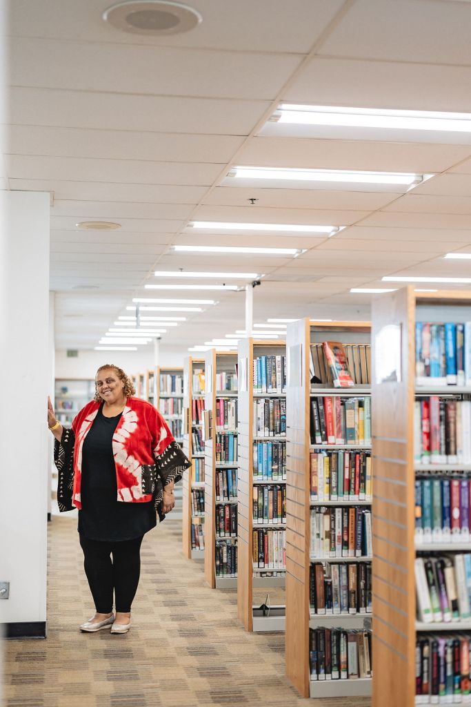 Deborah Anna Brown wearing a red and white patterned kimono-style jacket stands and smiles next to a bookshelf at the end of a brightly lit library aisle filled with books.