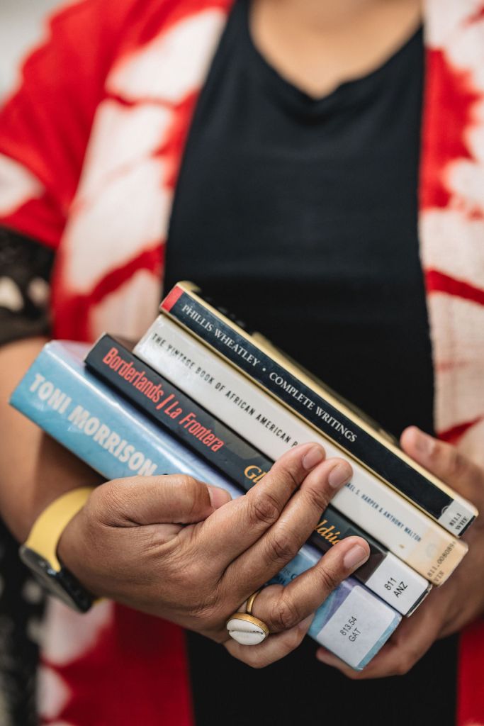 Deborah Anna Brown wearing a red and white patterned kimono style jacket with dark shirt holds four books, including works by Toni Morrison, Gloria Anzaldúa, and Phillis Wheatley. Only their hands and torso are visible.