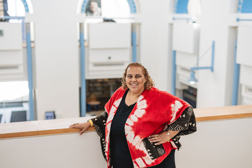 Deborah Anna Brown smiling indoors at a library, leaning on a wooden railing. She wears a red and white patterned kimono-style jacket over a black top and silver shoes. The background shows white walls and arches.