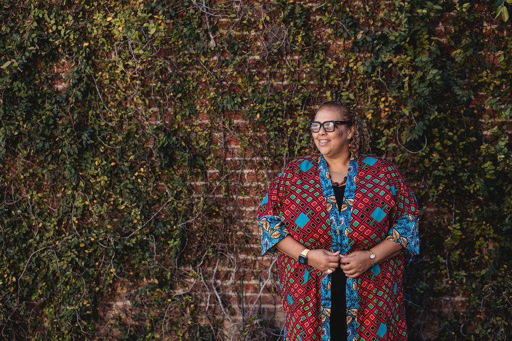 Deborah Anna Brown wearing glasses and a colorful patterned kimono-style jacket stands smiling, looking off into the distance. She is in front of a brick wall covered with green vines.