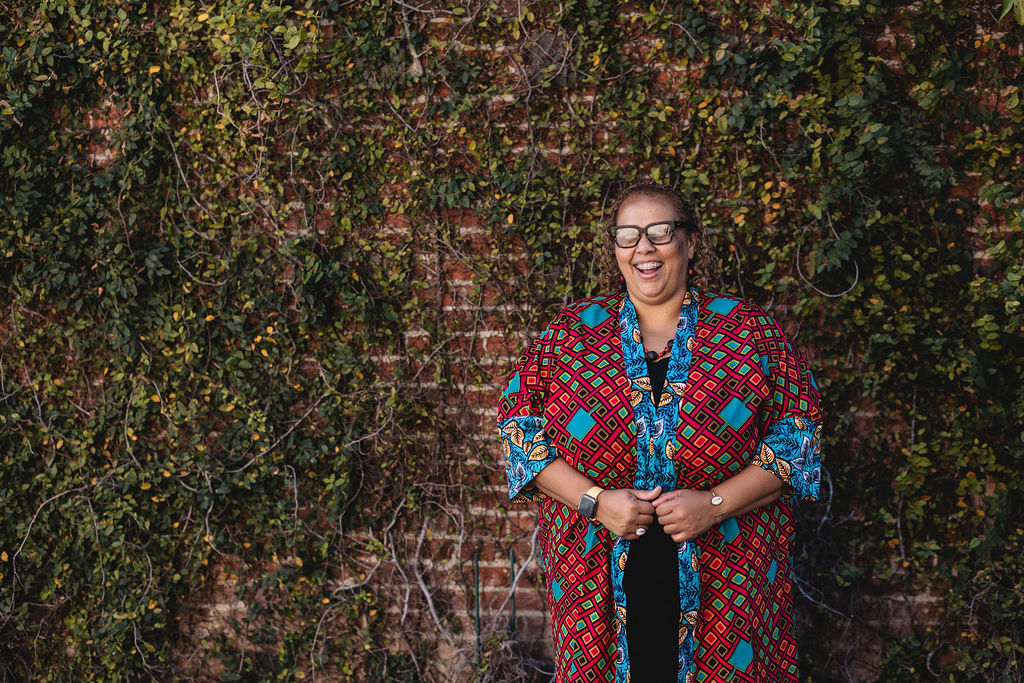 Deborah Anna Brown wearing glasses and a colorful patterned kimono-style jacket laughing in front of a brick wall covered in green vines.