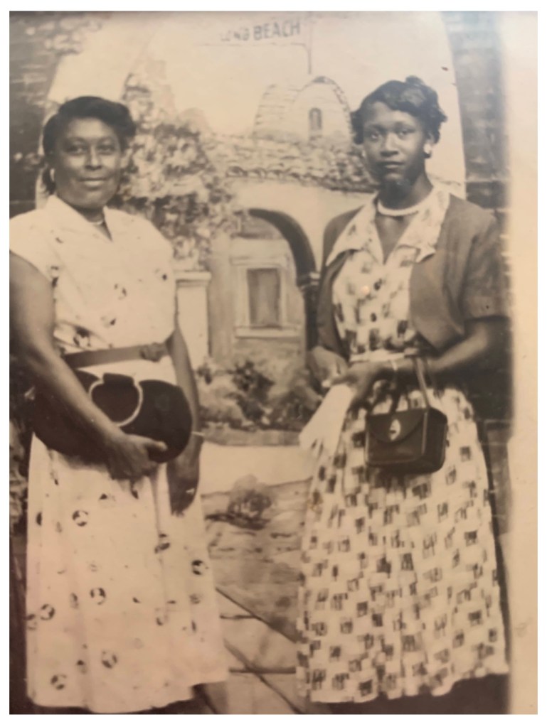 Two women in patterned dresses stand side by side, each holding a handbag, posing in front of a painted backdrop that includes the words LONG BEACH and depicts a domed building and plants.