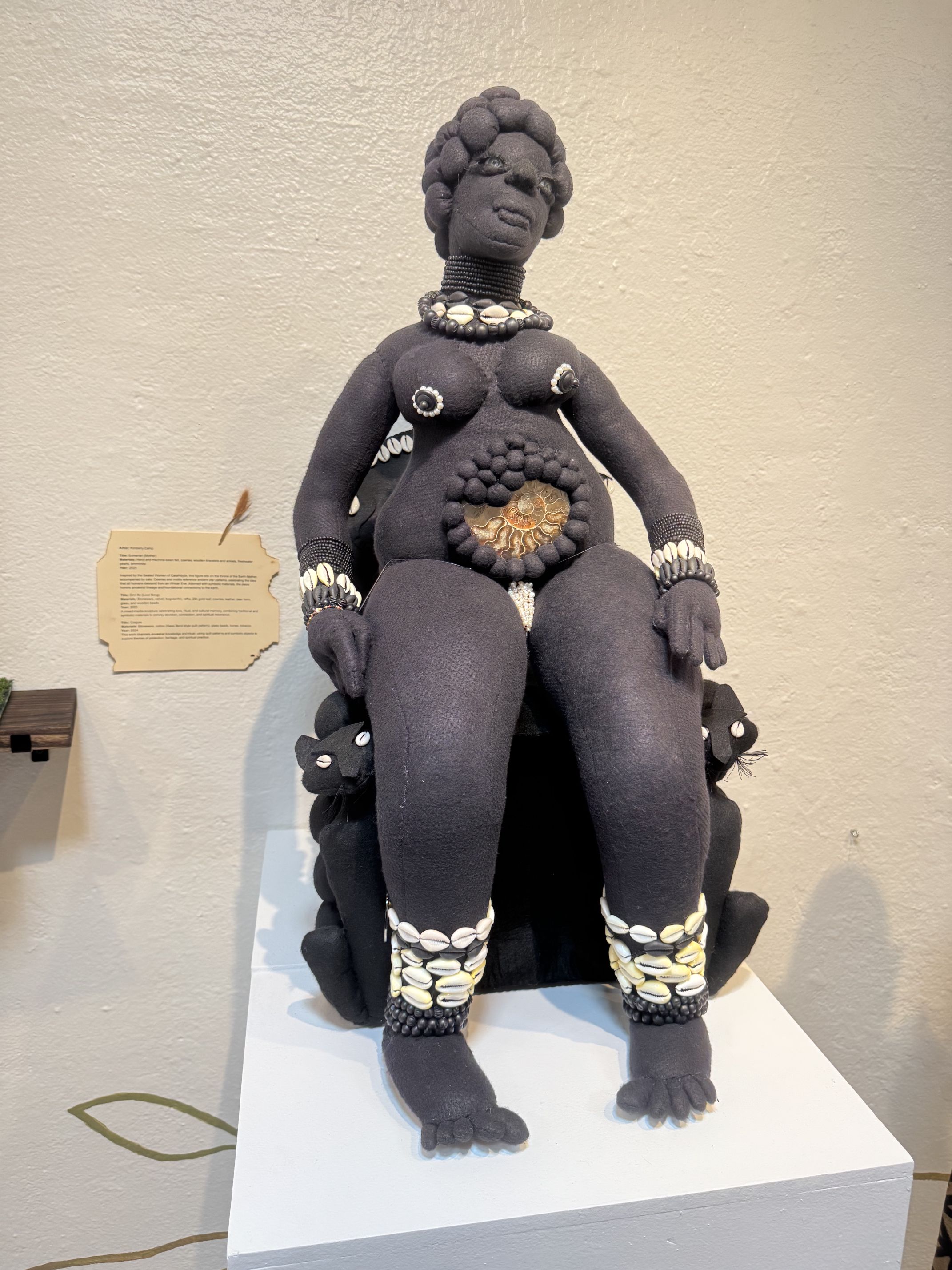 A textile sculpture of a seated woman adorned with shells, featuring braided hair and jewelry, and sitting on a black chair with cat figures on each side. The sculpture is displayed on a white pedestal against a beige wall.